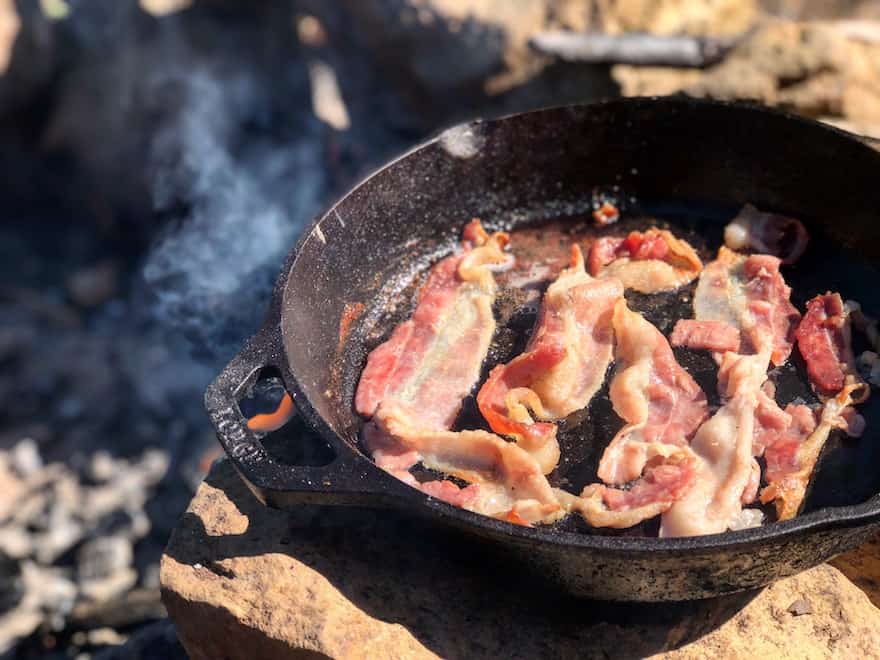 Bacon sizzling in a cast iron frying pan by a campfire