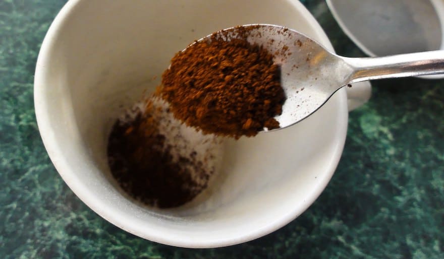 A spoon pours instant coffee crystals into an empty cup