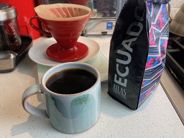 A cup of brewed coffee next to a bag of beans from Ecuador and a Hario V60 pour over cone in the background