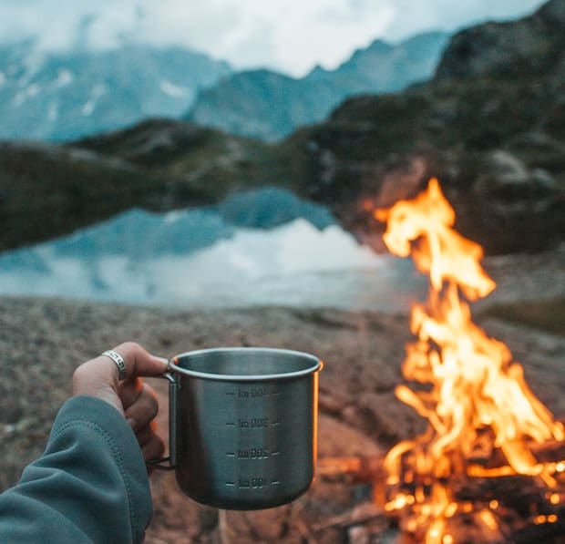 Hand holding up a tin coffee cup by a campfire