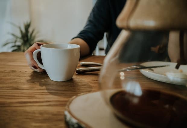 A person reaching for a cup of coffee with a Chemex carafe in the foreground