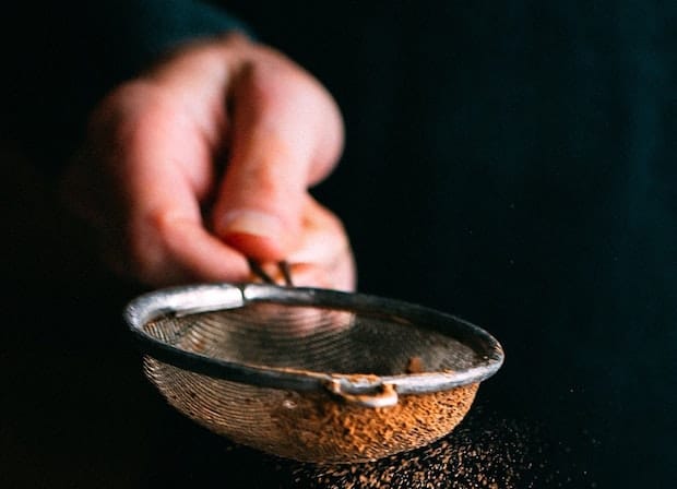 Hand holding a mesh sieve containing brown powder