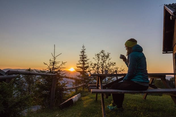 woman drinking coffee on a picnic table outside a cabin in the mountains at sunrise