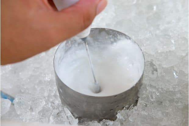 Hand using a handheld milk frother to make cold foam in a steel jug immersed in a bed of ice