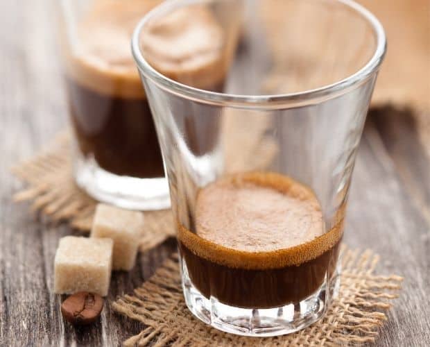 Small amount of espresso in the bottom of a shot glass, with a more full shot glass in the background