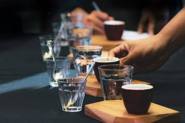 Man makes a list of coffee notes in the background while another tastes coffee from cups lined up on a table