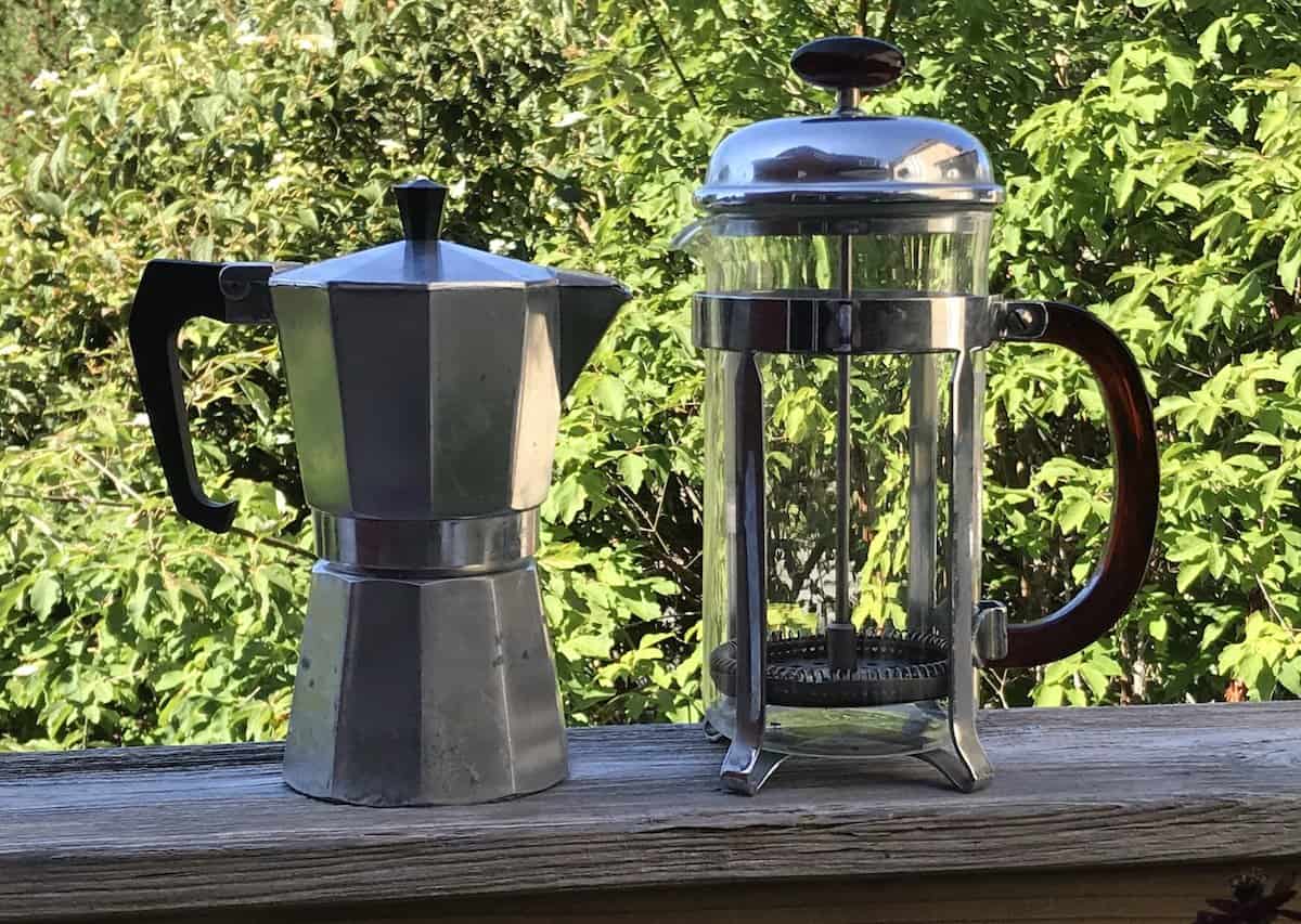 A moka pot and a French press side by side on the railing of a deck