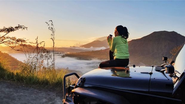 Woman sitting on vehicle watching sunrise and drinking coffee after using the best battery-powered coffee maker