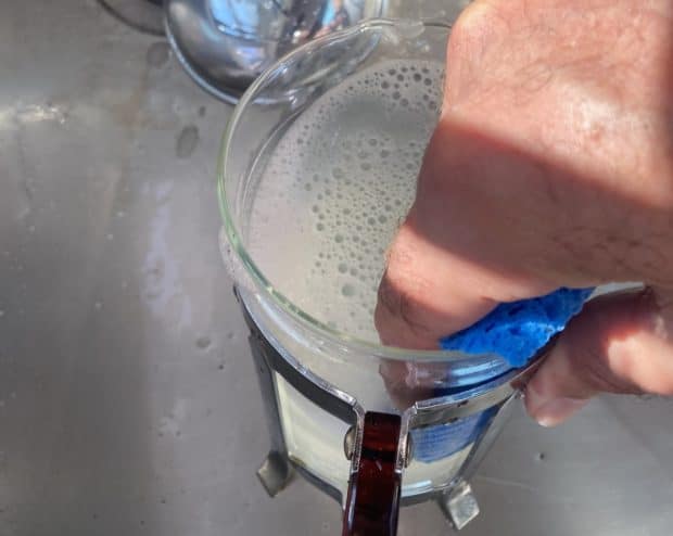 A hand washes a glass French press with a sponge and soapy water