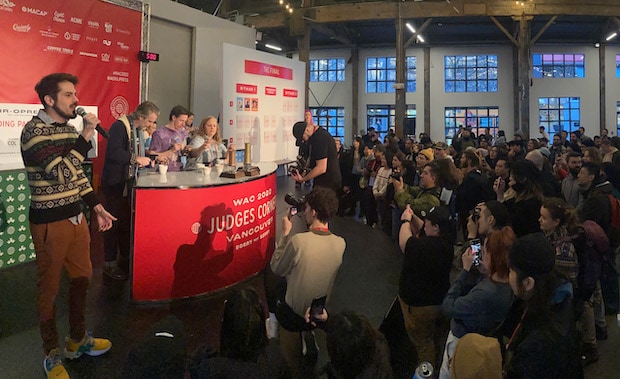 Judges taste coffee on a stage while a crowd awaits their verdict.