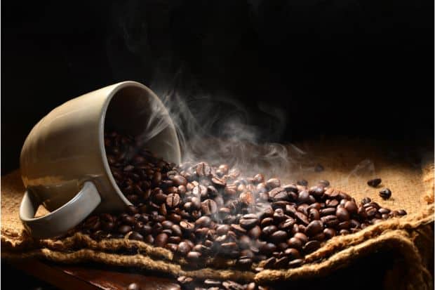 Coffee mug on its side with dark roast coffee beans spilling out amid a plume of smoke