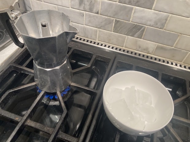 A bowl of ice cubes on a stove near a moka pot that is heating on a burner