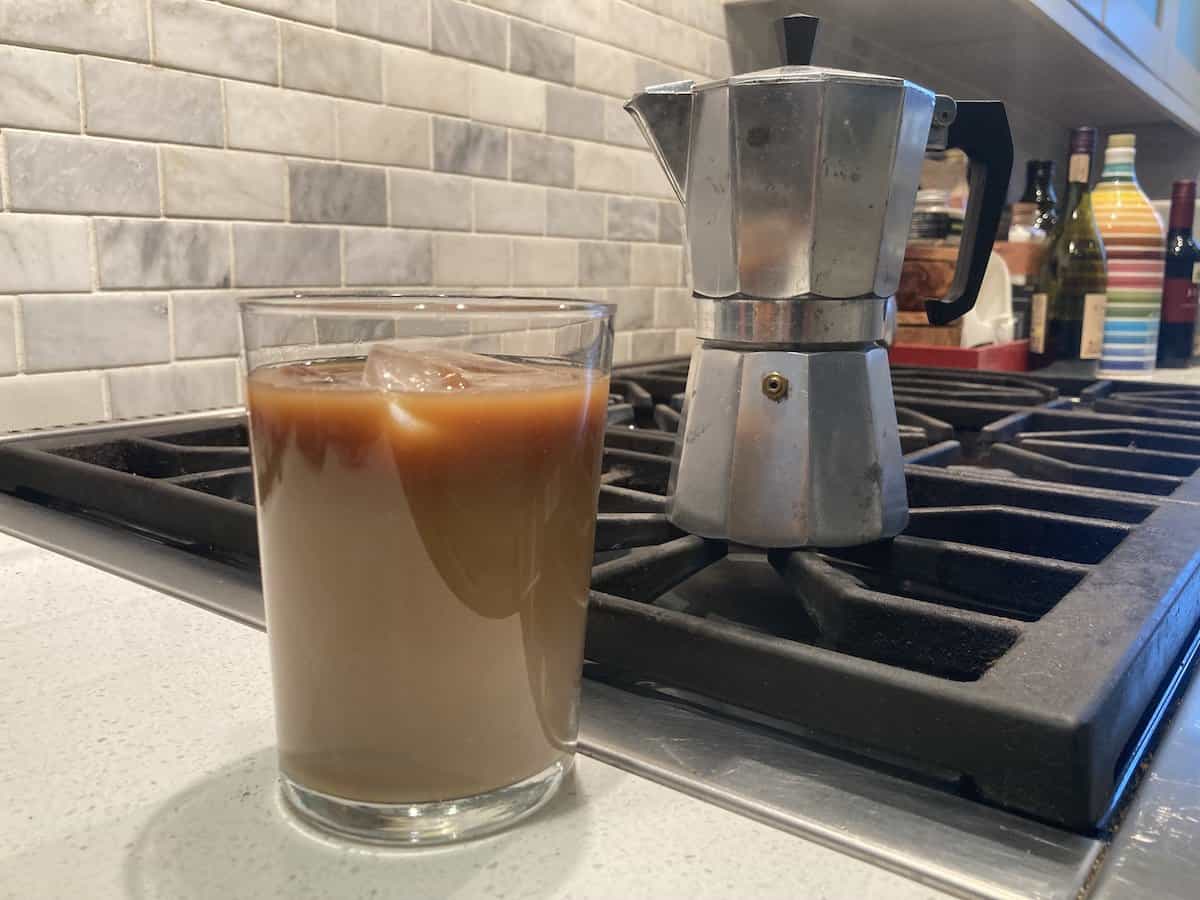 Glass of iced coffee in foreground with moka pot on stove in background