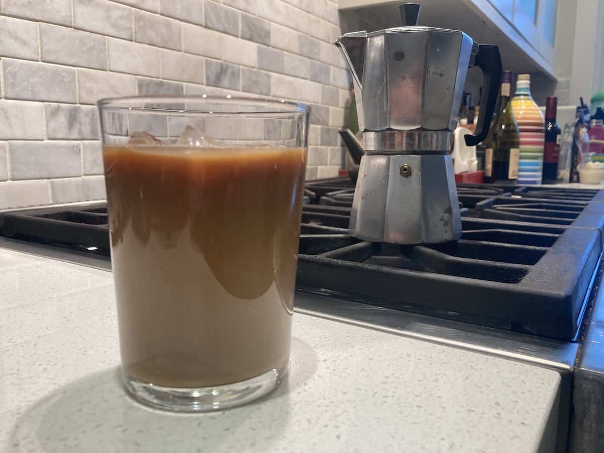 A glass of iced coffee in the foreground with a moka pot on the stove in the background
