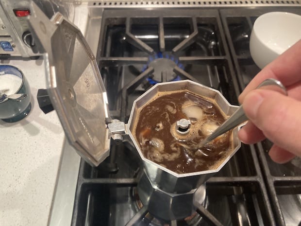 Hand with spoon stirs coffee and ice in upper chamber of a moka pot