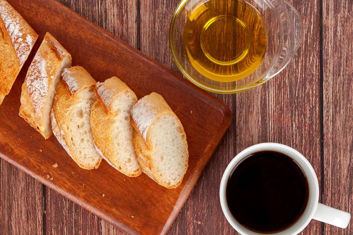 Sliced baguettes on a cutting board next to a carafe of olive oil and a cup of coffee