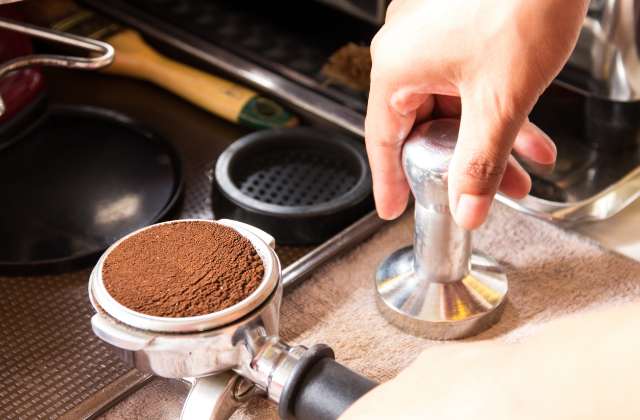 A portafilter filled with finely ground coffee and a hand getting ready to compress it with a tamper